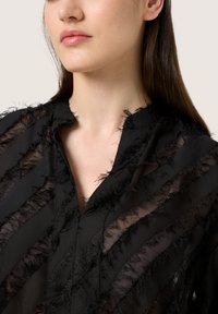 Blusa nera con un mix di tessuto trasparente e tessuto a trama, caratterizzata da strisce frangiate verticali e scollatura a V. Design testurizzato e a strati.