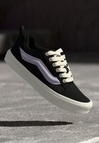 Svart sneaker med hvite snørebånd, lilla sidestropp og teksturert stoff. Har en flat gummisåle med avrundet tå.