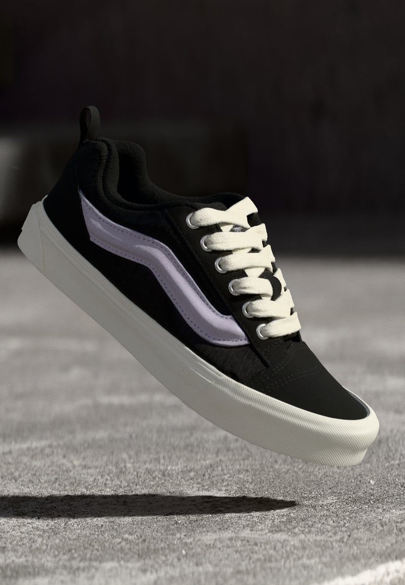 Svart sneaker med hvite snørebånd, lilla sidestropp og teksturert stoff. Har en flat gummisåle med avrundet tå.