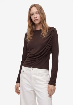 Femme aux longs cheveux blonds portant un haut moulant marron foncé à manches longues avec fronces latérales et un pantalon blanc taille haute sur fond blanc.