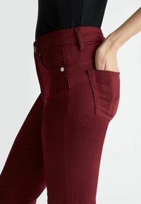 Bordowe jeansy o wąskim kroju, z boczną kieszenią i srebrnymi detalami. Wykonane z elastycznego materiału dla wygody i swobody ruchów.