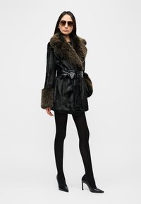 Μαύρο παλτό από συνθετική γούνα με φαρδιά γούνινη γιακά, ζώνη στη μέση και φλορά σαλιάρα στα μανίκια, συνδυασμένο με μαύρες καλσόν και ψηλοτάκουνες γόβες στιλέτο.