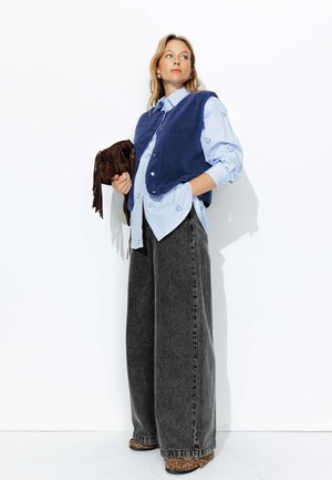 Pantalons en jean à jambes larges, chemise à rayures bleues avec broderies florales, gilet en polaire bleu et sac à franges marron. Chaussures à imprimé léopard.