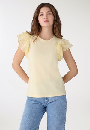 Giovane donna con capelli castano ondulati che indossa una canotta giallo pallido senza maniche con volant traforati e jeans azzurri.