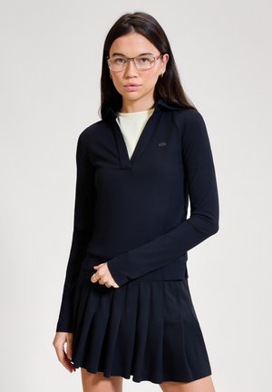 Jeune femme aux cheveux longs et portant des lunettes, vêtue d'une chemise noire à manches longues avec col par-dessus un haut blanc et d'une jupe plissée noire.