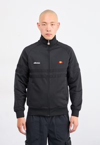 Mężczyzna ubrany w czarną kurtkę z zamkiem błyskawicznym z logo ellesse i czarne spodnie, stojący na tle białym.