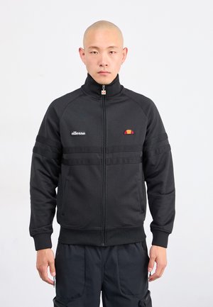 Man wearing black zip-up jacket with ellesse logo and black pants, standing against plain white background.