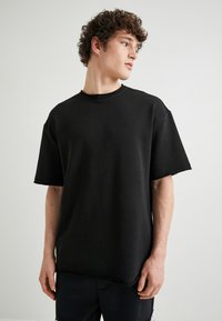Schwarzes, übergroßes T-Shirt aus weicher Baumwolle. Mit rundem Hals, kurzen Ärmeln und einem unfertigen Saum sowie einer leicht strukturierten Oberfläche.