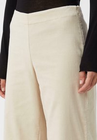 Beżowe spodnie w prążek z wysokim stanem, z szerokimi nogawkami, delikatnym pionowym prążkowaniem, zamkiem błyskawicznym z boku i gładką fakturą.