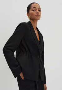 Zwarte blazer met een taillering, twee voorzakken en een enkele knoopsluiting. Gemaakt van een gladde stof met een op maat gemaakt ontwerp.