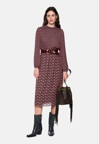 Langes burgunderrotes Kleid mit geometrischem Muster, gerafften Ärmeln und hohem Ausschnitt. Brauner Gürtel, schwarze Stiefeletten und schwarze Handtasche mit Fransen.