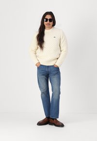 Cream knitted sweater with textured pattern, blue denim jeans, and brown leather loafers. Model wears black sunglasses and has long hair.