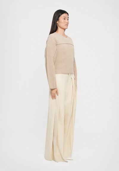 Maglione beige lavorato a maglia con scollatura ampia e maniche lunghe, abbinato a pantaloni plissettati color crema a vita alta. Dettaglio in tessuto testurizzato.