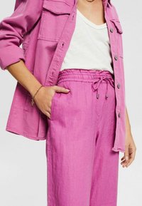 Femme portant une chemise boutonnée rose vif avec poches poitrine, pantalon à cordon de serrage assorti rose, haut blanc et bracelet en or, main dans la poche.