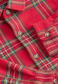 Camicia di flanella rossa con motivo a quadri con accenti verdi e gialli, chiusura con bottoni e tessuto morbido. I bottoni sono di colore marrone chiaro.