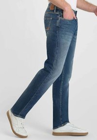 Jeans en denim bleu foncé avec une coupe droite, des coutures visibles et une étiquette en cuir à l'arrière de la taille. Portés avec des baskets blanches.