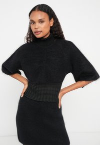 Μαύρο πλεκτό πουλόβερ με ψηλό ριχτό γιακά, κοντά μανίκια και υφασμάτινη ζώνη που εφαρμόζει. Μαλακό, τριχωτό υλικό με κομψή σιλουέτα.
