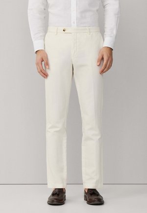 Man draagt slim-fit off-white broek, wit overhemd met knopen en donkerbruine loafers met kwastjes, staand tegen een effen achtergrond.