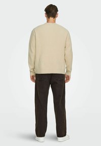 Beige gerippter Pullover mit Rundhalsausschnitt, langem Ärmel und lockerer Passform, kombiniert mit dunklen, baggy Jeans und weißen Sneakern.