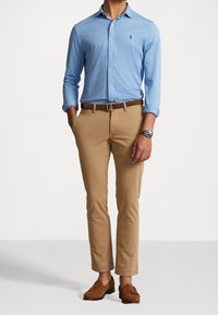 Jasnoniebieska koszula na guziki, khaki spodnie, brązowy skórzany pasek i brązowe mokasyny. Charakterystyczny zegarek na nadgarstku i podwinięte rękawy.