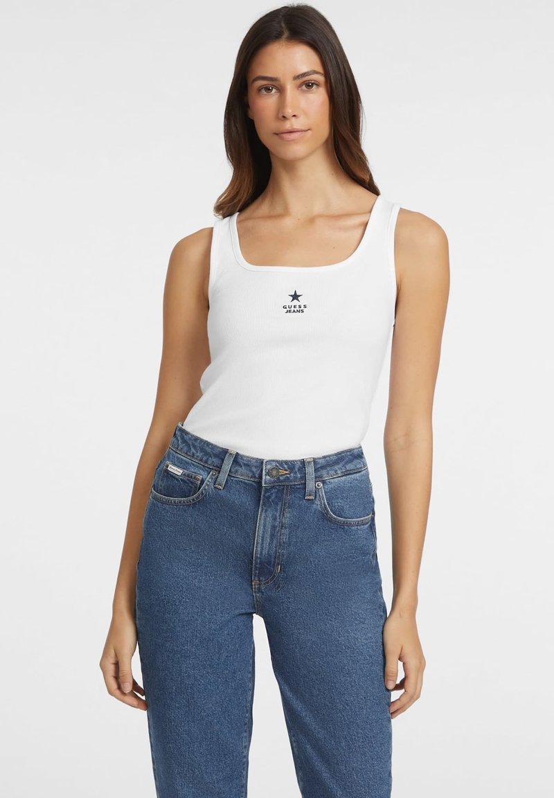 Valge sooniline tops, millel on ruudukujuline kaelus, keskosas must tähend ja "GUESS JEANS" logo, koos kõrge vöökohtadega siniste teksadega.