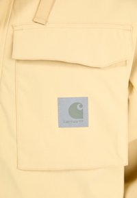 Camicia di tessuto giallo con taschino frontale e una toppa grigia con un logo circolare. Texture liscia con dettagli di cucitura visibili.