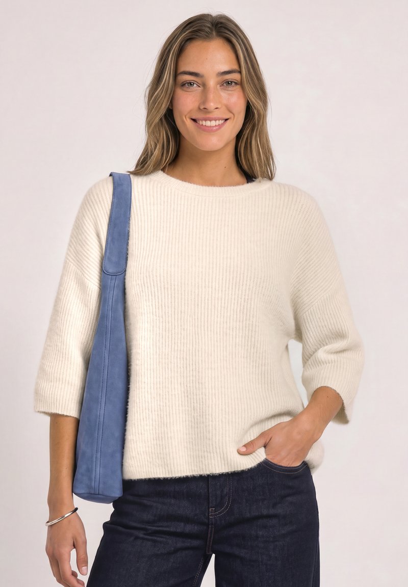 Femme souriante portant un pull blanc et un jean foncé, tenant un sac à épaule bleu avec la main gauche dans la poche et le bras droit détendu sur le côté.