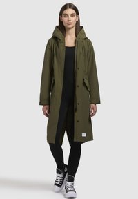 Parka longue vert olive avec une capuche, des boutons à l'avant, deux poches latérales et des cordons de serrage réglables. Portée sur une tenue noire et des baskets.