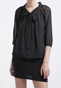 Kvinna klädd i en genomskinlig svart blus med en framknutning och trekvartsärmar, som är matchad med en figursydd svart kjol.