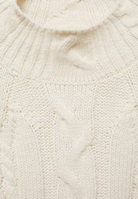 Suéter de color crema de punto con cuello acanalado y patrones de punto en cable, que muestra una suave y texturizada tela de lana.