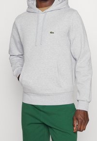 Uomo che indossa una felpa leggera grigia con tasca a marsupio e logo verde di coccodrillo, abbinata a pantaloni verdi, su uno sfondo semplice.