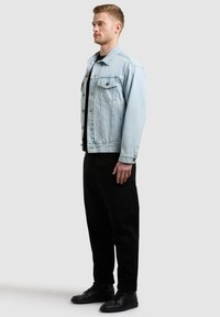 Jeune homme de profil portant une veste en jean bleu clair, un t-shirt noir, un pantalon noir et des baskets noires sur un fond uni.