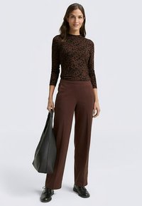 Braunes langärmliges Oberteil mit Leopardenmuster, kombiniert mit hoch taillierter brauner Hose und schwarzen Schuhen. Trägt eine große schwarze Tragetasche.