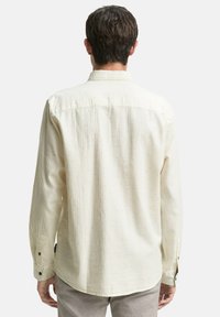 Mann mit kurzen braunen Haaren, der ein elfenbeinfarbenes langärmliges Hemd mit Knöpfen trägt, von hinten vor einem schlichten Hintergrund zu sehen.