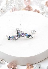 Silverfärgade örhänge med en böjd design prydd med flera fasetterade ädelstenar i lila, blå, grön och klar färg, åtskilda av en vit bakgrund.