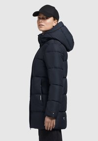 Navy puffer coat with a hood, featuring a quilted design, side pockets, and a shorter hem. Paired with a black cap. Smooth texture.