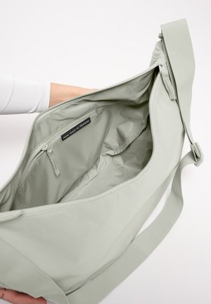 Borsa a tracolla grigio chiaro tenuta aperta, che mostra l'interno vuoto con tasca con cerniera ed etichetta di stoffa con la scritta "FROM TRASH TO TREASURE."