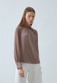 Long-sleeve blouse in taupe fabric features a keyhole neckline, pleated detailing, and fitted cuffs. Smooth texture with a slight sheen.