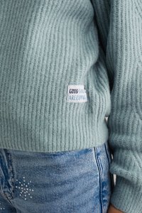 Šviesiai mėlyna, raukinėta megztinė su etikete "GERI DALYKAI ATĖJA", derinama su mėlynomis džinsinėmis kelnėmis, turinčiomis dekoratyvių detalių.