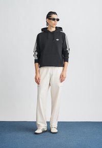 Juodas džemperis su baltomis juostelėmis ant rankovių ir logotipu, derinamas su kreminėmis siaurėjančiomis kelnėmis ir baltais sportbačiais. Modelis dėvi tamsius akinius nuo saulės.