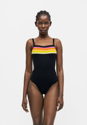 Femme portant un maillot de bain une pièce noir avec des rayures horizontales rose, orange et jaune sur la poitrine, debout devant un fond uni.