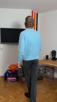 Pulover pufos albastru deschis, cu croială lejeră, asortat cu blugi gri închis. Fundalul include un televizor, o lampă portocalie și diverse obiecte de decor.