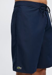 Σκούρα μπλε μαγιό βερμούδα με πράσινο λογότυπο κροκόδειλου στο αριστερό πόδι και μαύρο κορδόνι δεμένο στη μέση.