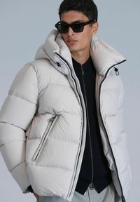 Doudoune blanche avec col haut et capuche, dotée de poches zippées et d'une finition brillante, portée sur un sweat à zipper sombre.