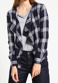 Navyblårandig kavaj med draperad front, långa ärmar och mjukt tyg, lager på lager över en grå skjorta och mörka jeans.