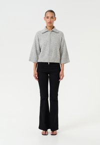 Giacca grigia, taglio corto con zip e colletto, maniche larghe. Indossata sopra jeans neri svasati. Tessuto liscio, design minimalista.