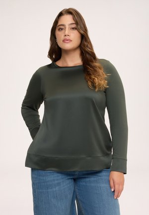 Kvinde med langt bølget hår iført en mørkegrøn langærmet top og blå jeans, stående med den ene hånd bag ryggen.