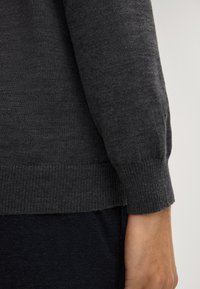 Pull gris avec poignets et ourlet côtelés, fabriqué en tissu tricoté doux. Texture lisse avec une coupe ajustée, montrant une main reposant sur le poignet.