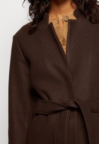 Cappotto di lana marrone con cintura indossato sopra un maglione beige a maglia con grandi bottoni, mostrato dalle spalle fino a metà torso.
