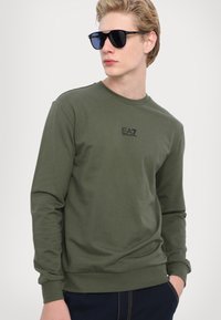 Olive zelená mikina s dlhým rukávom, s klasickým okrúhlym výstrihom a rebrovanými manžetami. Na hrudi sa nachádza malé čierne logo EA7. Hladký bavlnený materiál.